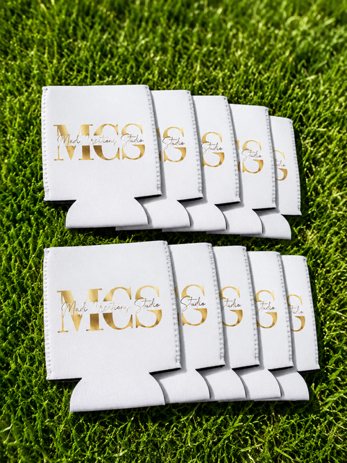 Diez koozies de neopreno blancos impresos con el logo dorado de MCS sobre el pasto