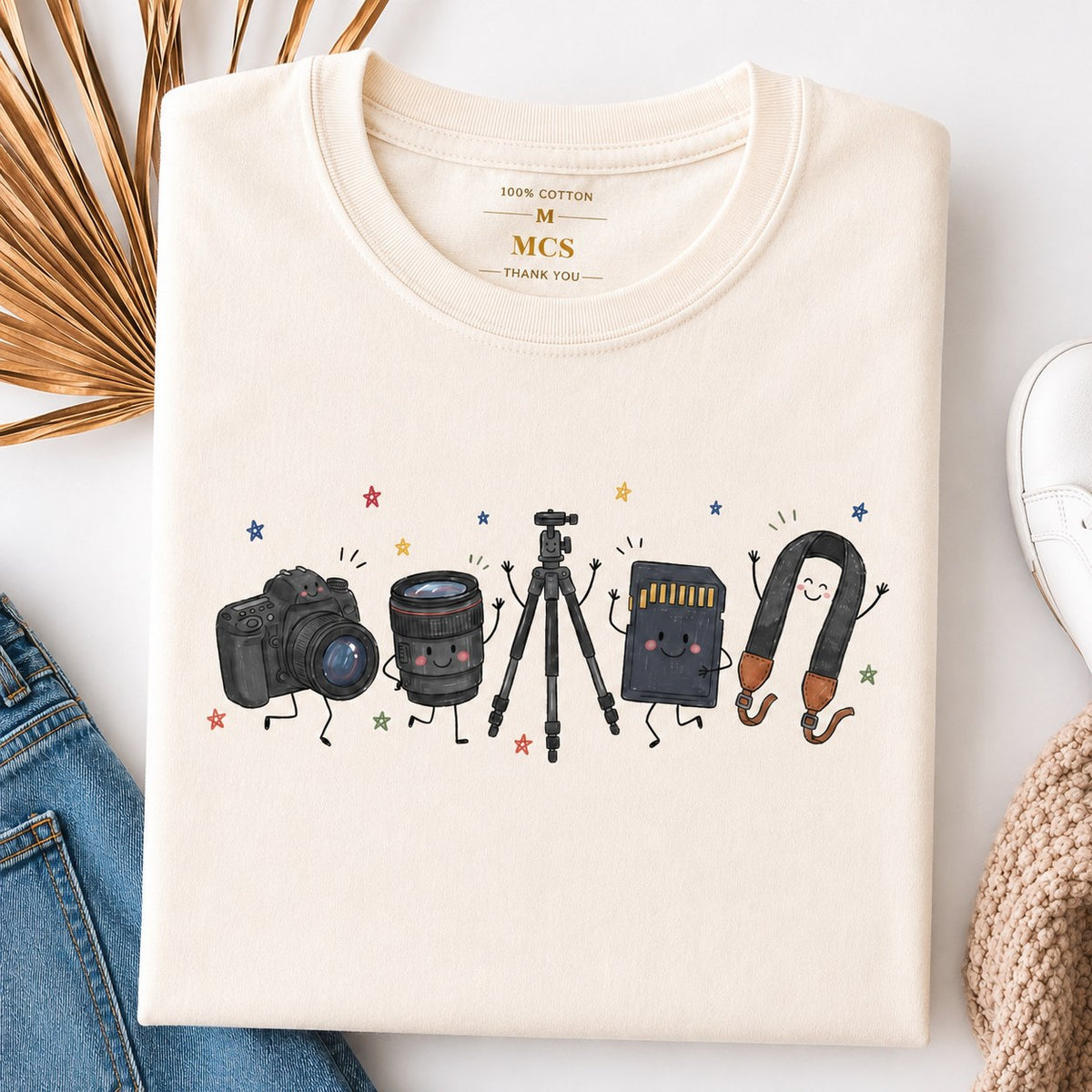 Cream t-shirt with cartoon camera, lens, tripod, memory card and camera strap characters