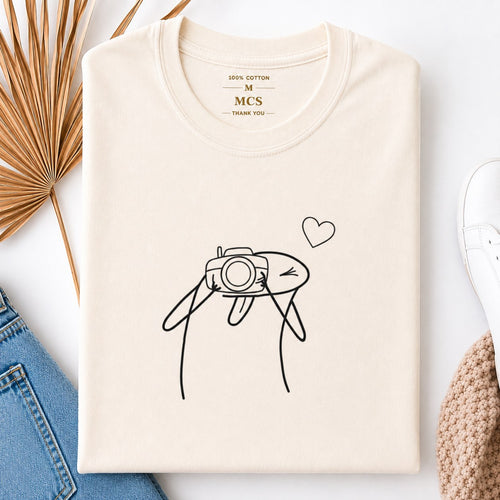 Cream t-shirt with minimalist line art of a character taking a photo with a camera and a little heart