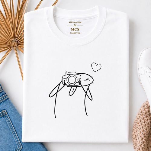 White t-shirt with minimalist line art of a character taking a photo with a camera and a little heart