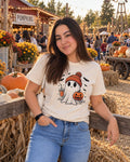 Woman wearing a cream t-shirt printed with a ghost in an orange beanie holding an iced drink and a mini pumpkin