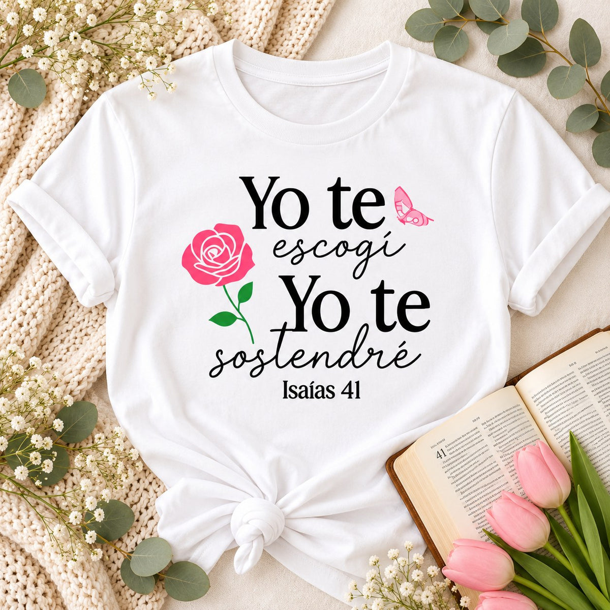 White t-shirt flat lay printed with Yo te escogí, Yo te sostendré, a rose, a butterfly and Isaías 41, next to a Bible and tulips